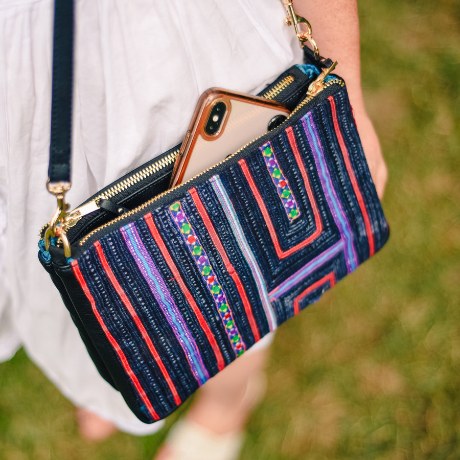 Photo of Woman in white dress walking outside wearing Rafi Nova Black Sapa Crossbody Bag. Woman carrying bag with Navy, red and purple fabric detailing. Phone sitting in pocket. 